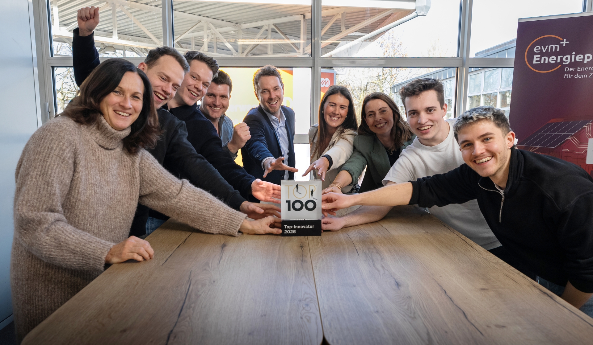 Ein Team der evm versammelt sich um einen Besprechungstisch und präsentiert gemeinsam den ‚Top 100‘‑Innovationspreis, der vor ihnen auf dem Tisch steht. Mehrere Personen strecken die Hände in Richtung der Auszeichnung aus und zeigen sichtbar Freude über den Erfolg.