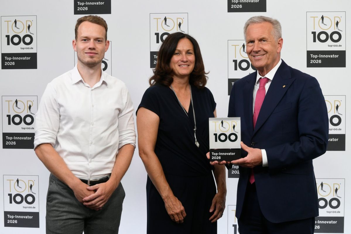 Top 200-Award Christian Wulff (rechts) überreicht den TOP 100‑Award an Julia Holly und Janos Justen von der evm. Die drei stehen vor einer TOP‑100‑Fotowand, während Wulff die Auszeichnung „Top‑Innovator 2026“ präsentiert.