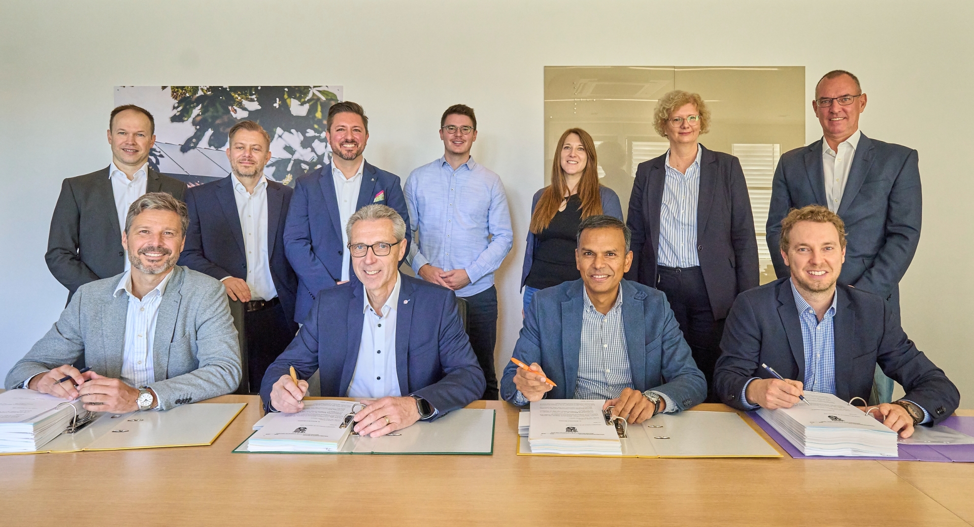 Vertreterinnen und Vertreter von evm und Iqony stehen und sitzen gemeinsam an einem Konferenztisch. In der ersten Reihe unterzeichnen Matthias Ohl, Jürgen Kirsch, Mithun Basu und Christoph Hesse die Verträge zur Gründung der Wärme Moselbogen GmbH, während weitere Beteiligte im Hintergrund für ein Gruppenfoto zusammenstehen.