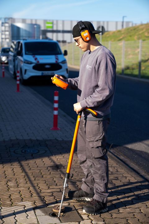 Ein Mitarbeiter mit Gehörschutz misst mit einem Gerät an einem offenen Gulli.