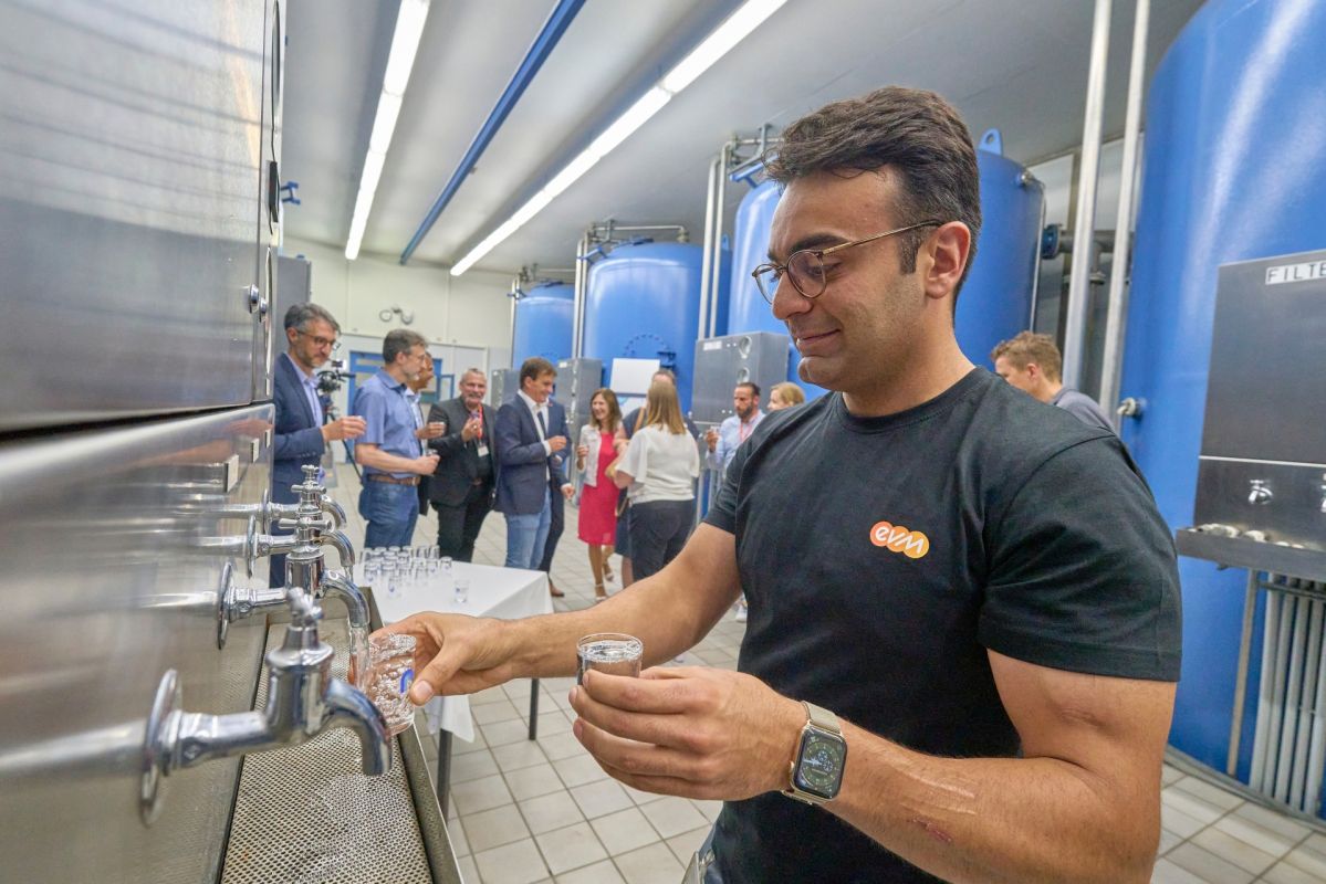 Ein Mann mit evm T-Shirt steht an einem Wasserspender und hält ein Wasserglas in der Hand. Er lächelt freundlich.