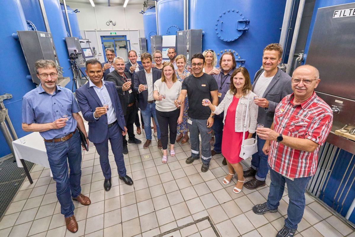 Eine Gruppe professionell gekleideter Menschen steht in einem Wasserwerk. Sie lächeln freundlich und halten Wassergläser in der Hand.
