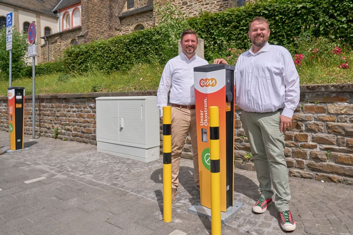 Zwei Männer stehen vor einer orangenen evm-Ladesäule und lächeln freundlich.