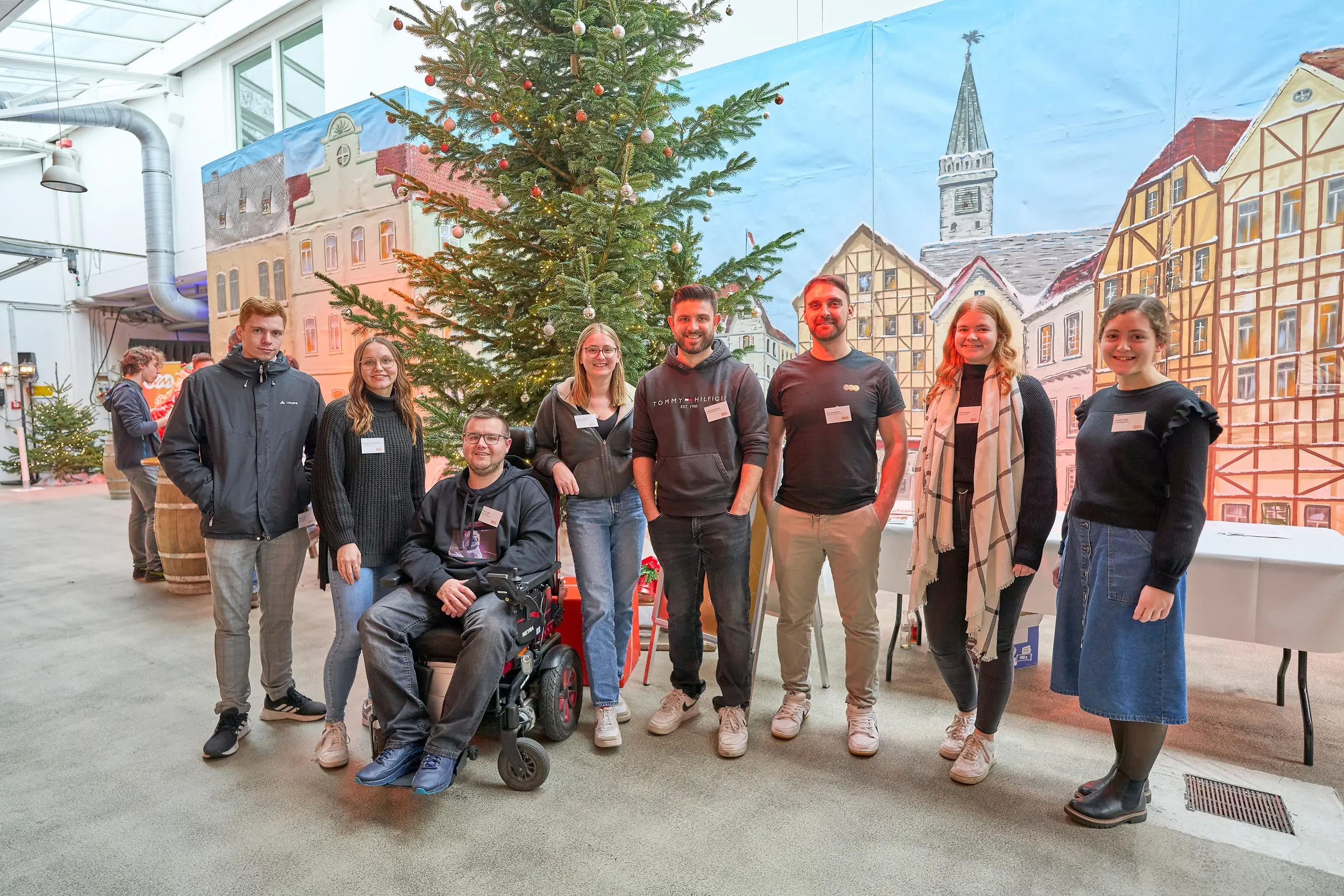 Eine Gruppe von acht Menschen steht in einer Halle vor einem Tannenbaum. Im Hintergrund sind Malereien von Fachwerkhäusern zu sehen. Jede Person trägt ein Namensschild.