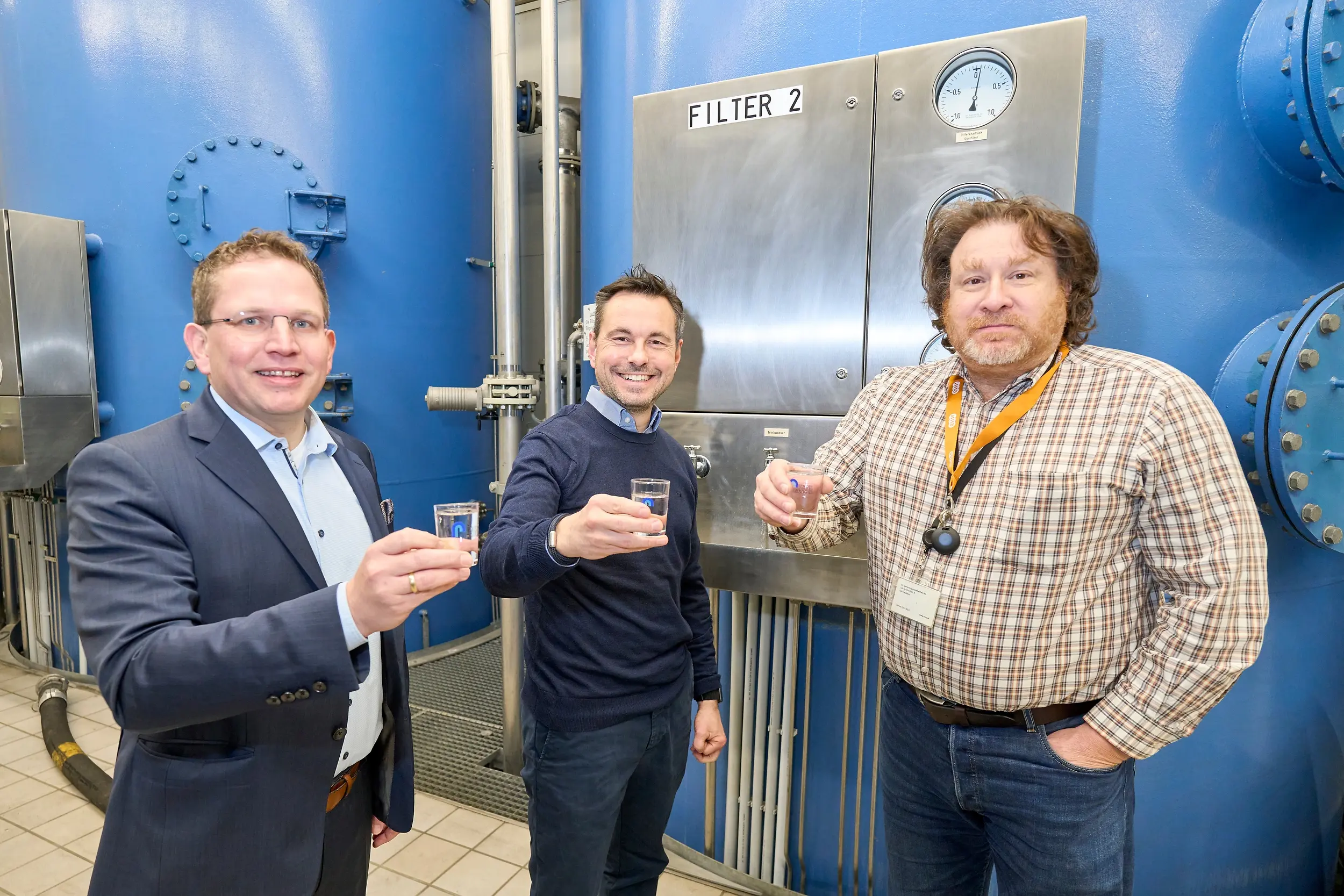 Drei Männer halten Gläser mit Wasser und stehen vor großen blauen Filtrationsanlagen. Einer trägt ein Namensschild und eine Stoppuhr. Oberhalb der Geräte befindet sich ein Schild: "FILTER 2".