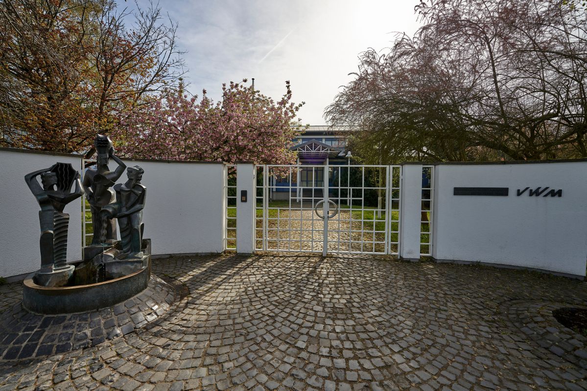 Der Eingang vom Wasserwerk Oberwerth. Eine weiße Wand mit einem Tor, daneben ein kleiner Brunnen.