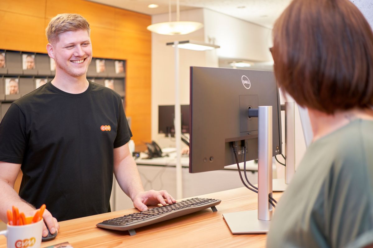 Ein Mann trägt ein schwarzes T-Shirt mit einem evm Logo und steht vor einem Computerbildschirm in einem Büro mit unscharfem Hintergrund. Er bedient eine Kundin.