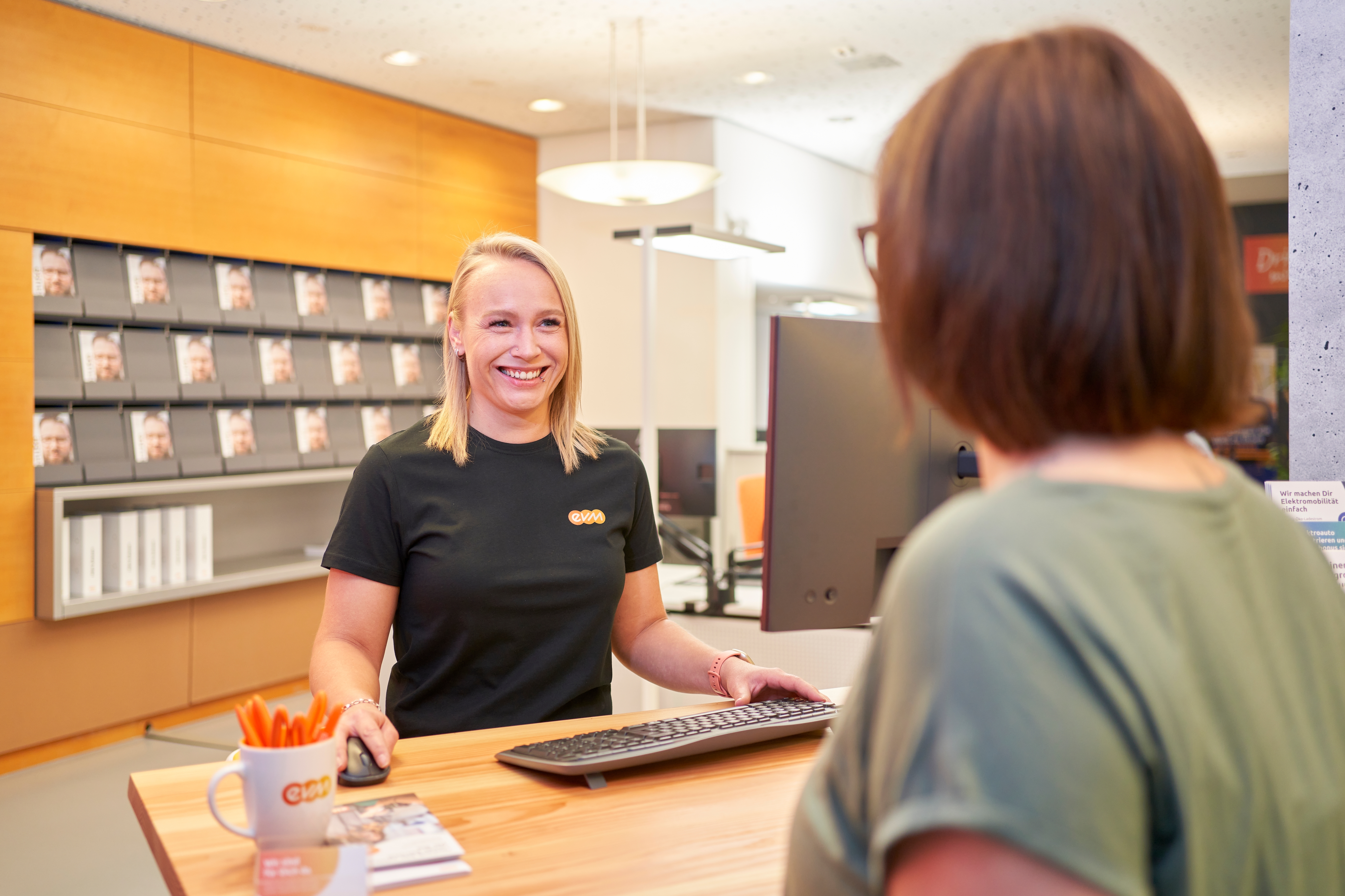 Eine lächelnde Frau steht an einem Schalter, bedient einen Computer und redet mit einer Kundin. Im Hintergrund sind Zeitschriftenregale und es gibt einen Becher mit Stiften, darauf steht "evm".