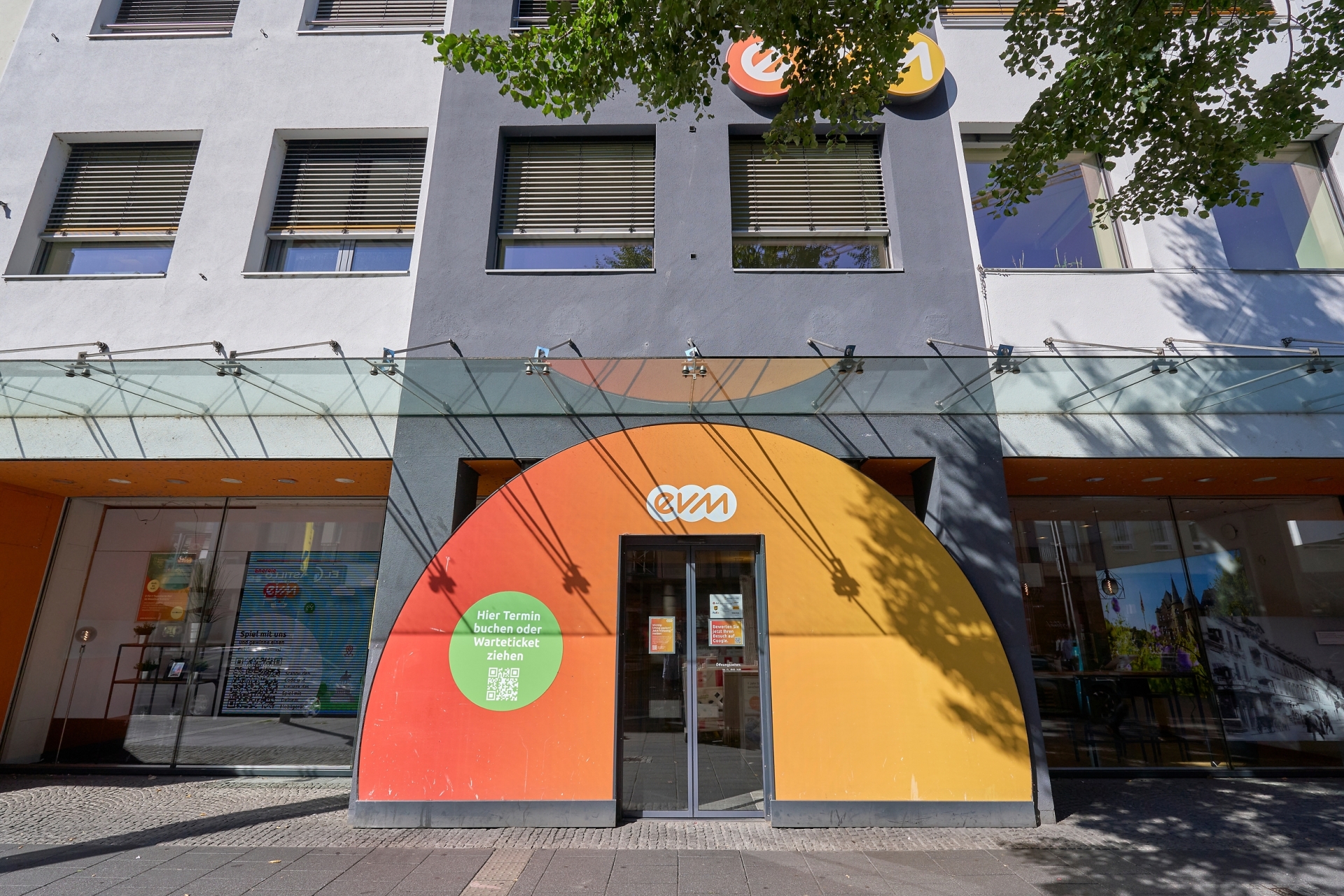 Eine halbkreisförmige orange-rote Wand mit einer Glastür in der Mitte und evm-Logo, vor einem Geschäft in einer Fußgängerzone.