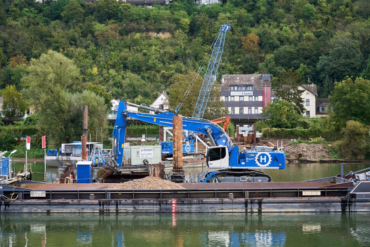 Ein blauer Bagger steht auf einem Ponton auf dem Wasser. Im Hintergrund sind Bäume und Häuser zu erkennen.