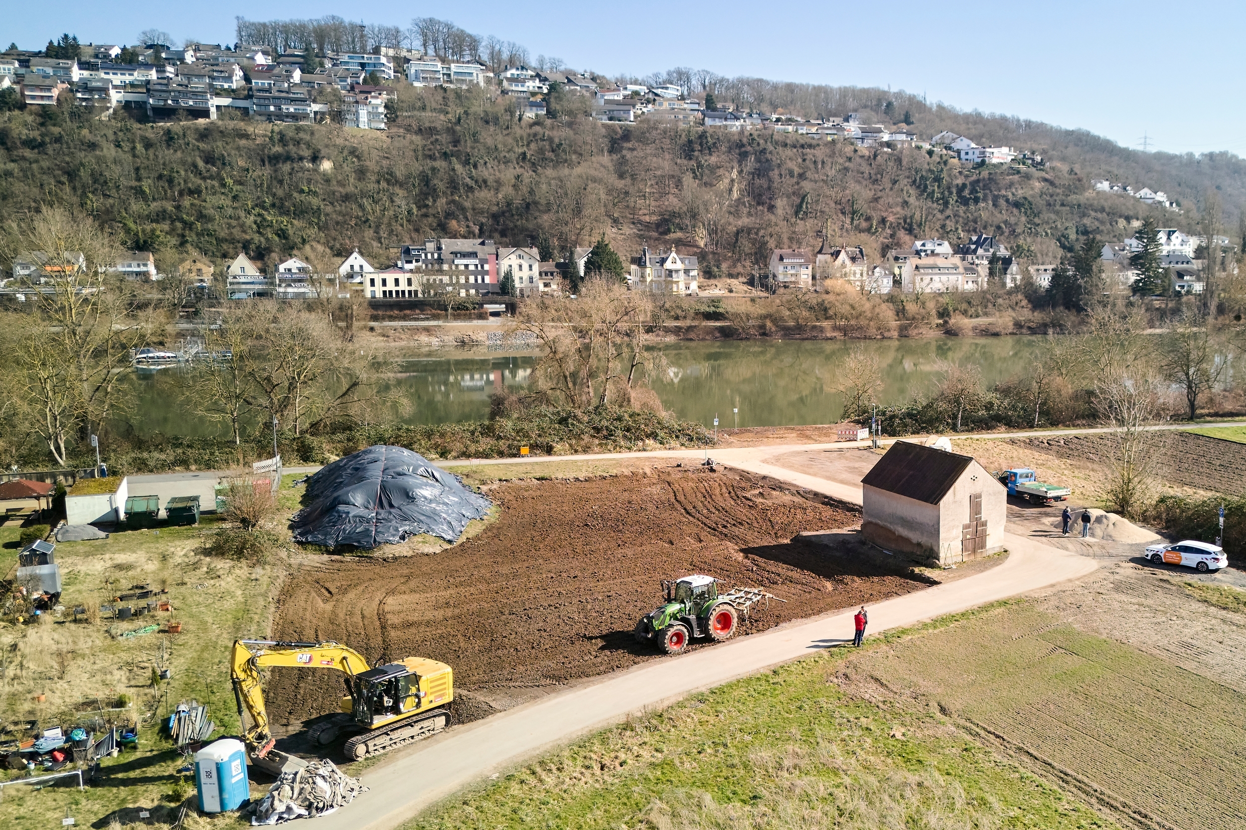Himmelblick auf eine Baustelle mit zwei Baggern. Im Hintergrund ist  ein Fluss sowie mehrere Häuser zu erkennen.