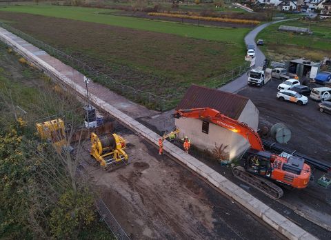 241120SDI1031 Himmelblick auf eine Baustelle mit einem orangenen Bagger und zwei Personen in orangener Schutzkleidung.