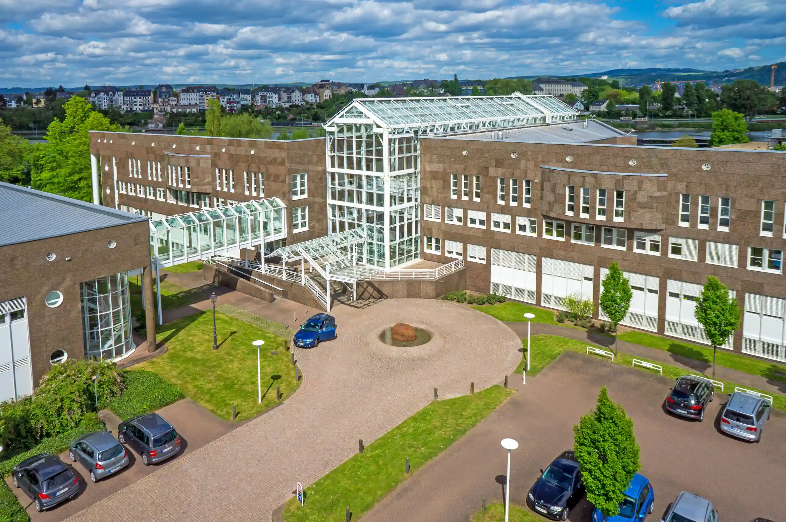 Ein mehrstöckiges Gebäude mit großen Fenstern und weißer Stahl-Glas-Eingangskonstruktion. Autos parken auf einem angrenzenden Platz, umgeben von gepflegten Grünflächen und Bäumen.