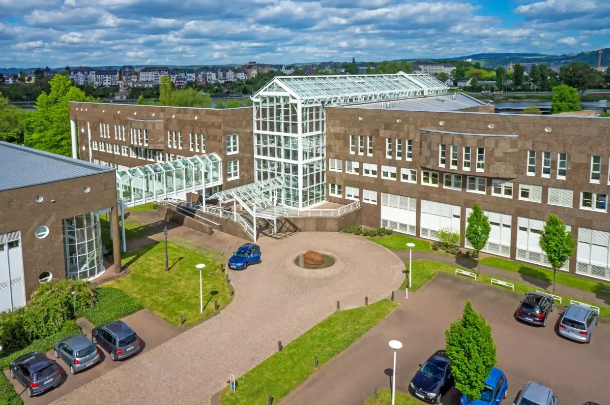 Ein mehrstöckiges Gebäude mit großen Fenstern und weißer Stahl-Glas-Eingangskonstruktion. Autos parken auf einem angrenzenden Platz, umgeben von gepflegten Grünflächen und Bäumen.