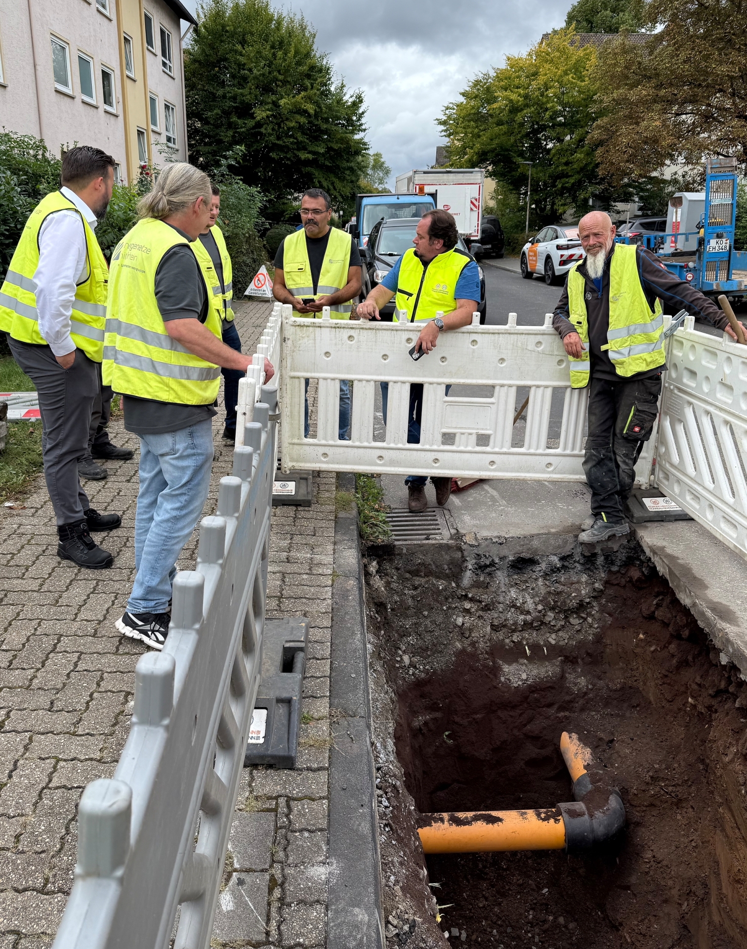 Mehrere Personen in gelben Schutzwesten stehen an einer Baustelle. Ein orangenes Rohr liegt offen in einem Graben.