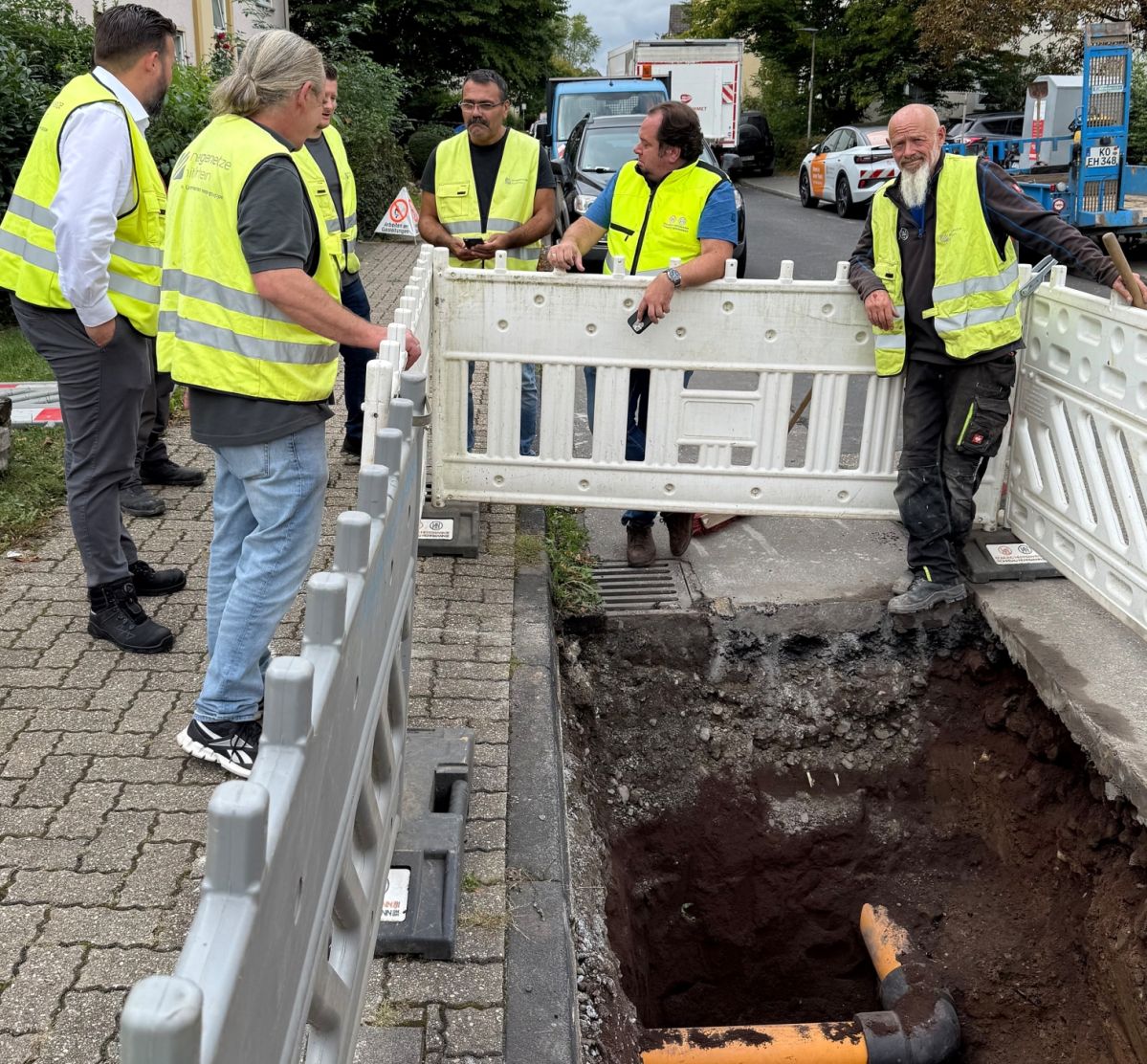 IMG_2526 Mehrere Personen in gelben Schutzwesten stehen an einer Baustelle. Ein orangenes Rohr liegt offen in einem Graben.