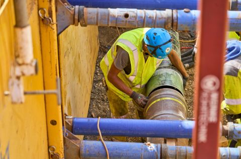 Reparatur Schaden: Gastransportleitung Andernach – Mayen / Straße: B 256 / Bereich Obere Löhr Straße Ein Arbeiter mit einer gelben Schutzweste und einem blauen Schutzhelm arbeitet an einem großen Rohr auf einer Baustelle.