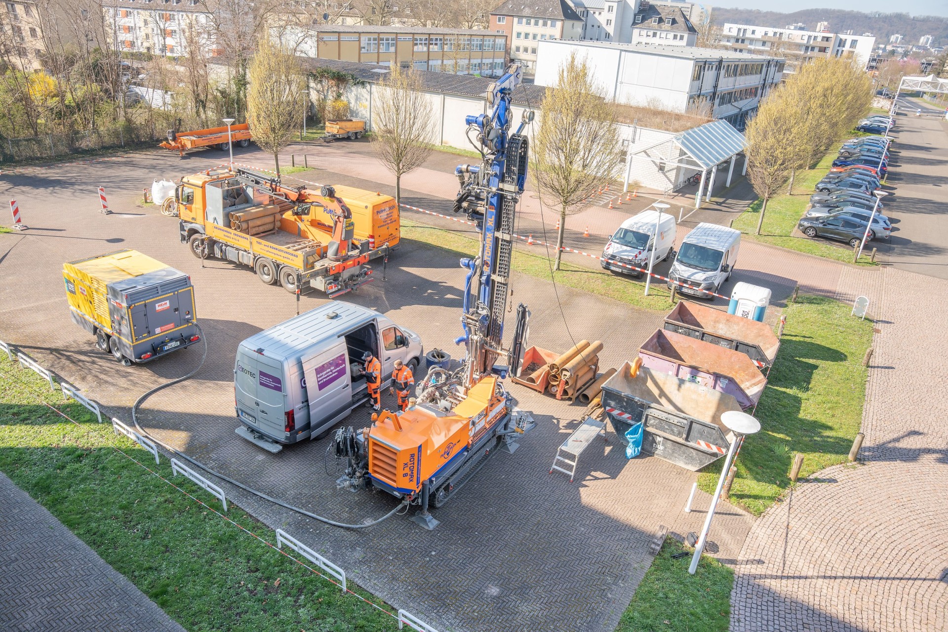 Himmelblick auf eine Baustelle einer Anodenanlage. Ein Bagger, ein Transporter und zwei Maschinen arbeiten auf einem Parkplatz. Zwei Personen in orangener Schutzkleidung stehen daneben.