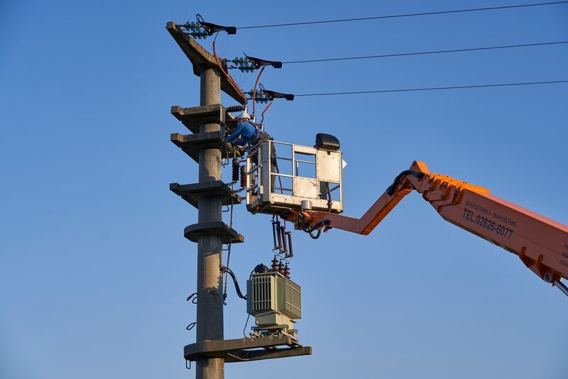 Strommasten werden überprüft Ein Techniker arbeitet in einem Hubwagen an einem Strommast. Der Mast trägt Kabel und einen Transformator. Klarer blauer Himmel bildet den Hintergrund.