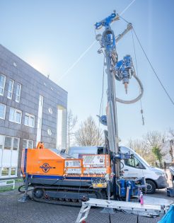 Ein orangener Bagger mit einem großen blauen Bohrer steht auf einem Parkplatzgelände.