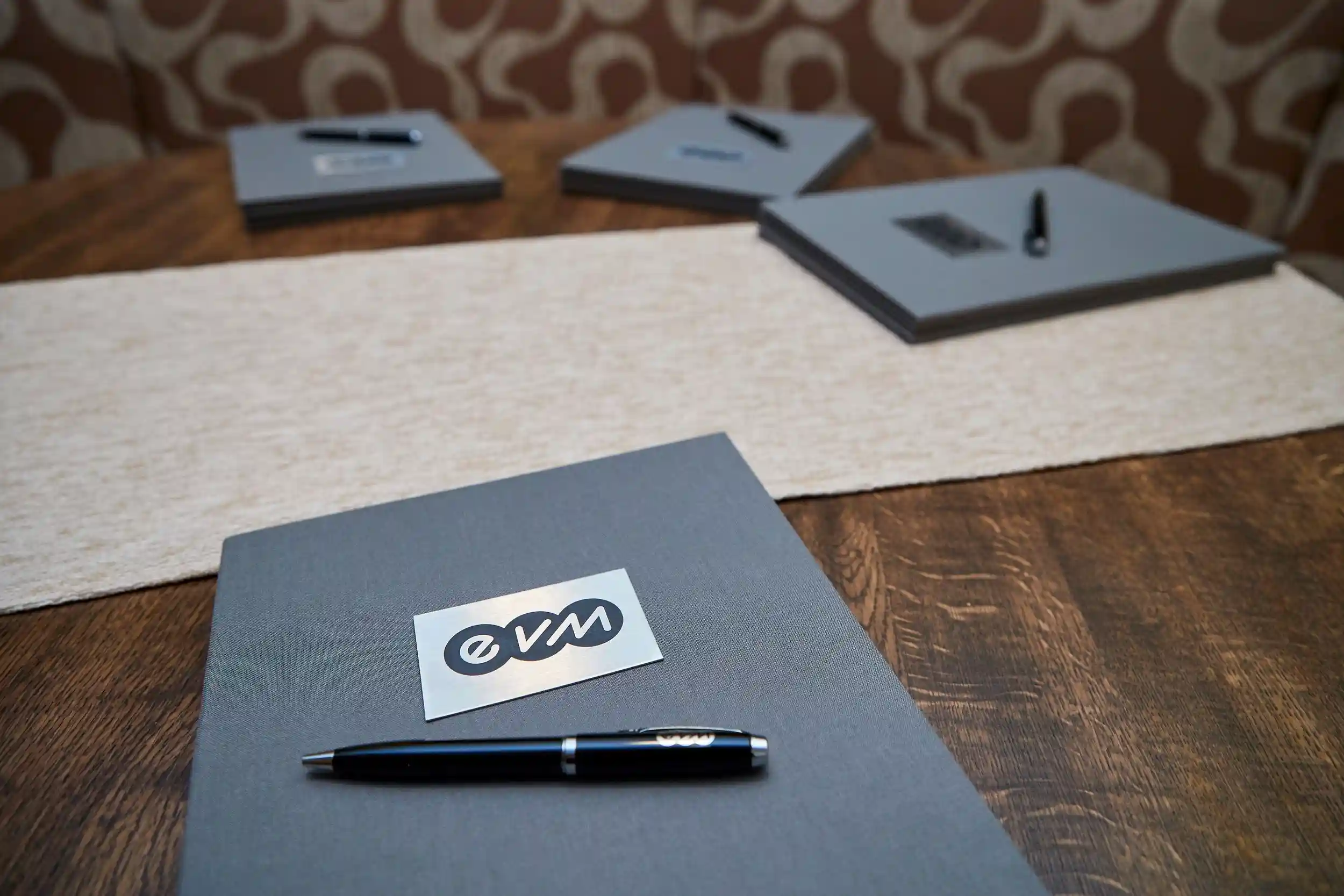 Ein schwarzer Kugelschreiber liegt auf einem grauen Notizbuch mit "evm"-Logo, platziert auf einem Holztisch. Mehrere identische Notizbücher und Stifte sind im Hintergrund verteilt.