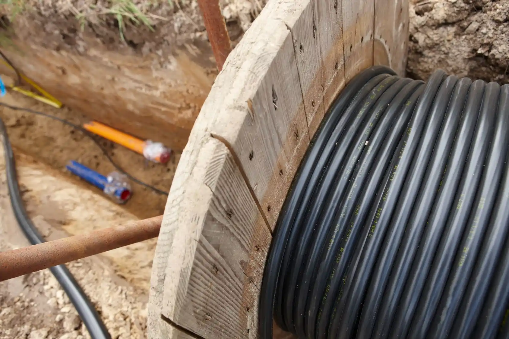 Eine große Kabeltrommel mit schwarzem Kabel wird in einem Erdgraben abgespult; im Hintergrund sind farbige Rohre zu sehen, die in den Boden verlegt werden.