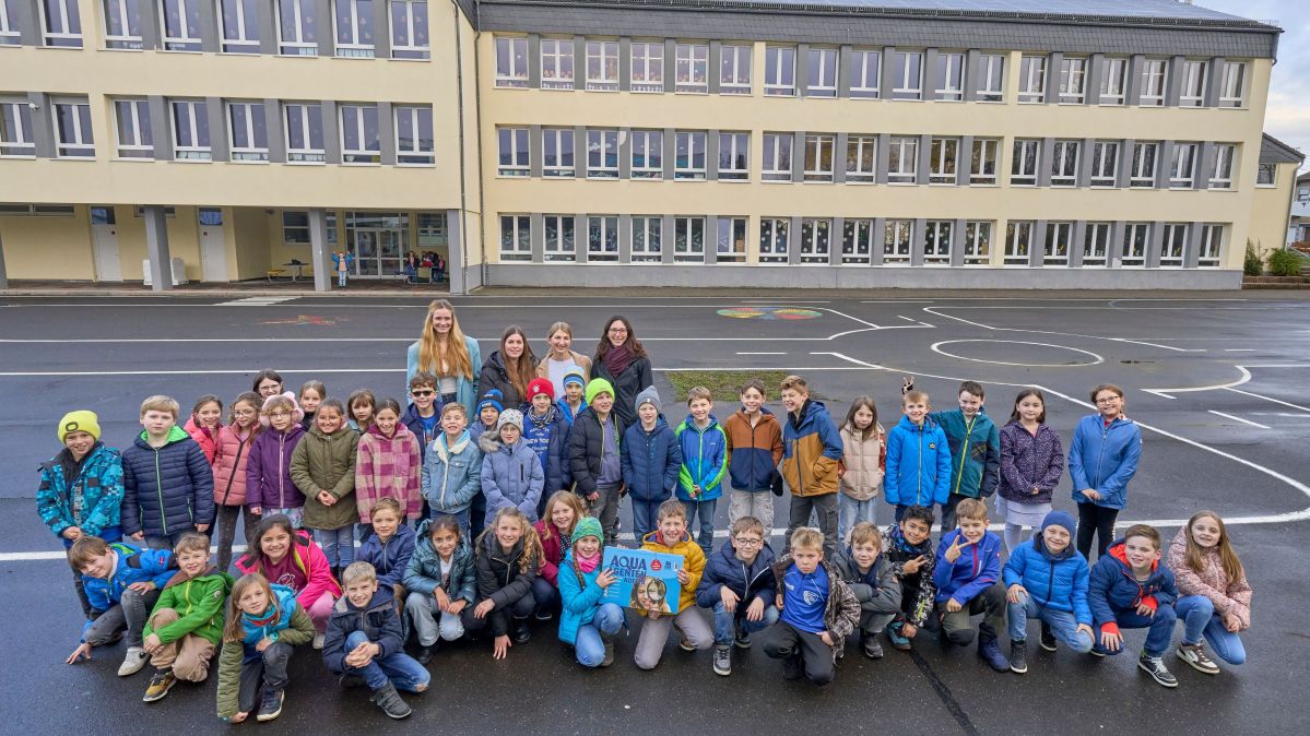 Eine Gruppe von Schulkindern stehen in einem Schulhof. Vier Frauen stehen dabei und lächeln freundlich.