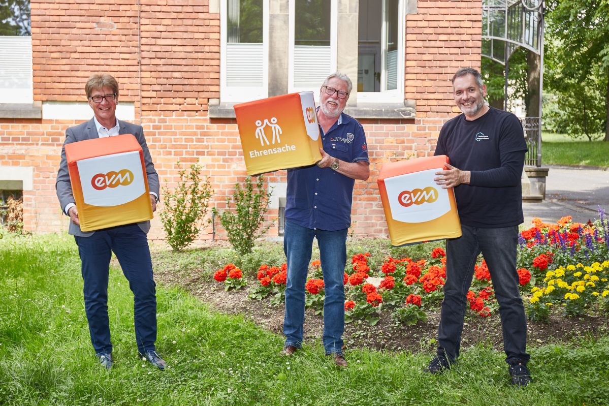 Drei Männer stehen auf einer Wiese und halten orangene Würfel mit der Aufschrift "evm" und "Ehrensache".