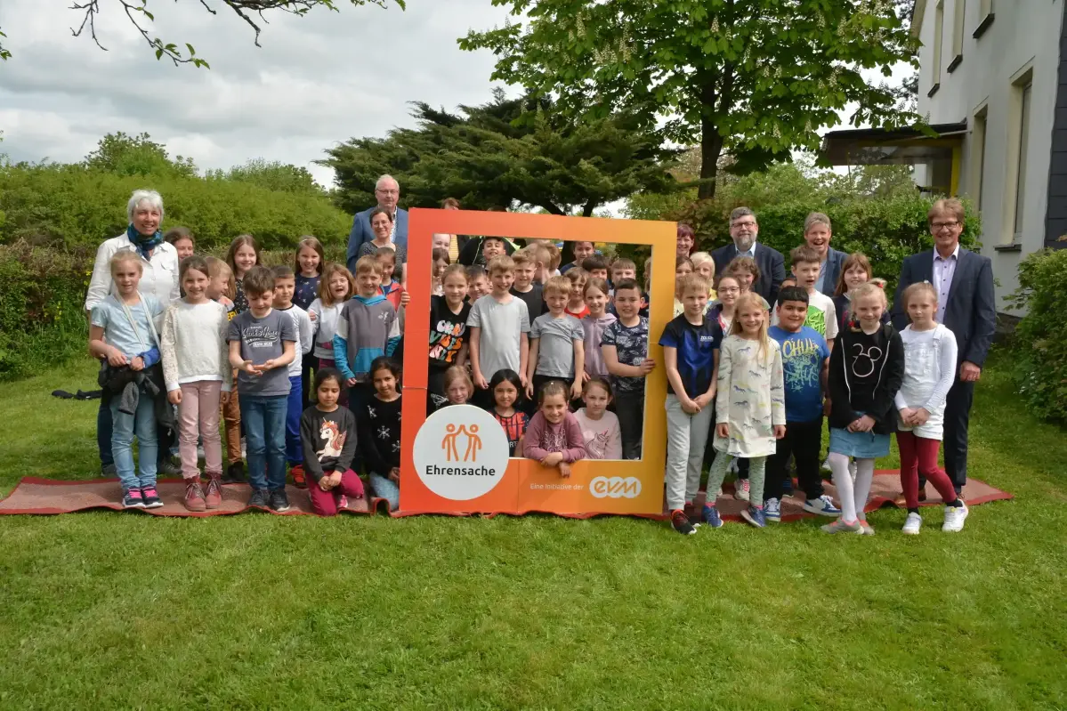 evm Ehrensache Cochem Eine Gruppe an Kindern und Erwachsenen stehen in einem Garten und halten einen orangenen Rahmen mit der Aufschrift "evm Ehrensache". Sie lächeln freundlich.