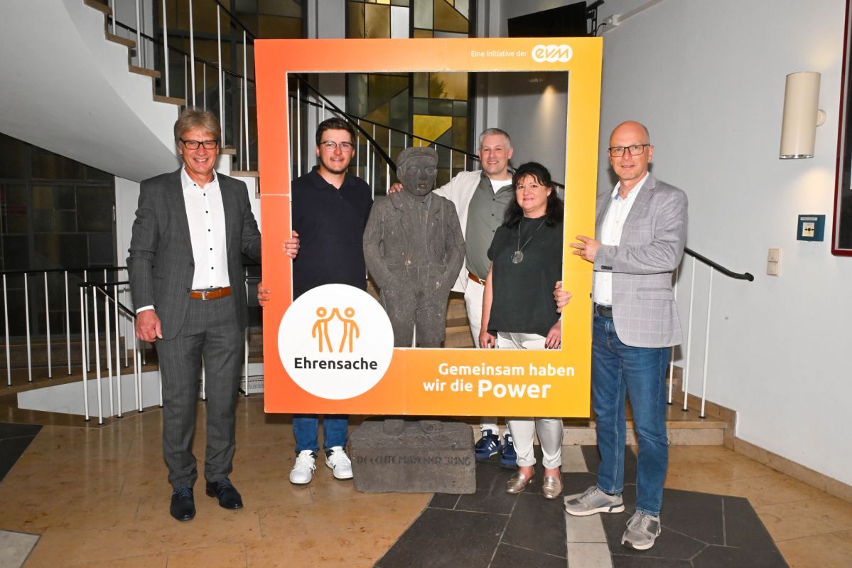 Vier Männer und eine Frau stehen vor einer Statue und halten einen Orangenen Rahmen. Auf dem Rahmen steht "evm Ehrensache". Sie lächeln freundlich und sind professionell gekleidet.