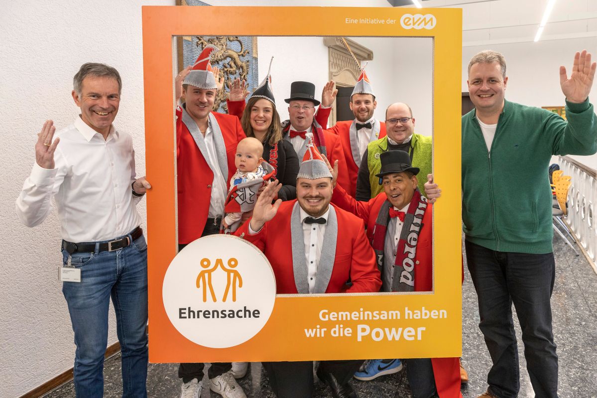 eVM Ehrensache Spende VG Rennerod_2 Eine Gruppe von Menschen, einige in festlichen Kostümen, lächelt und winkt innerhalb eines orangefarbenen Rahmens. Text im Rahmen: „Ehrensache“ und „Gemeinsam haben wir die Power“.