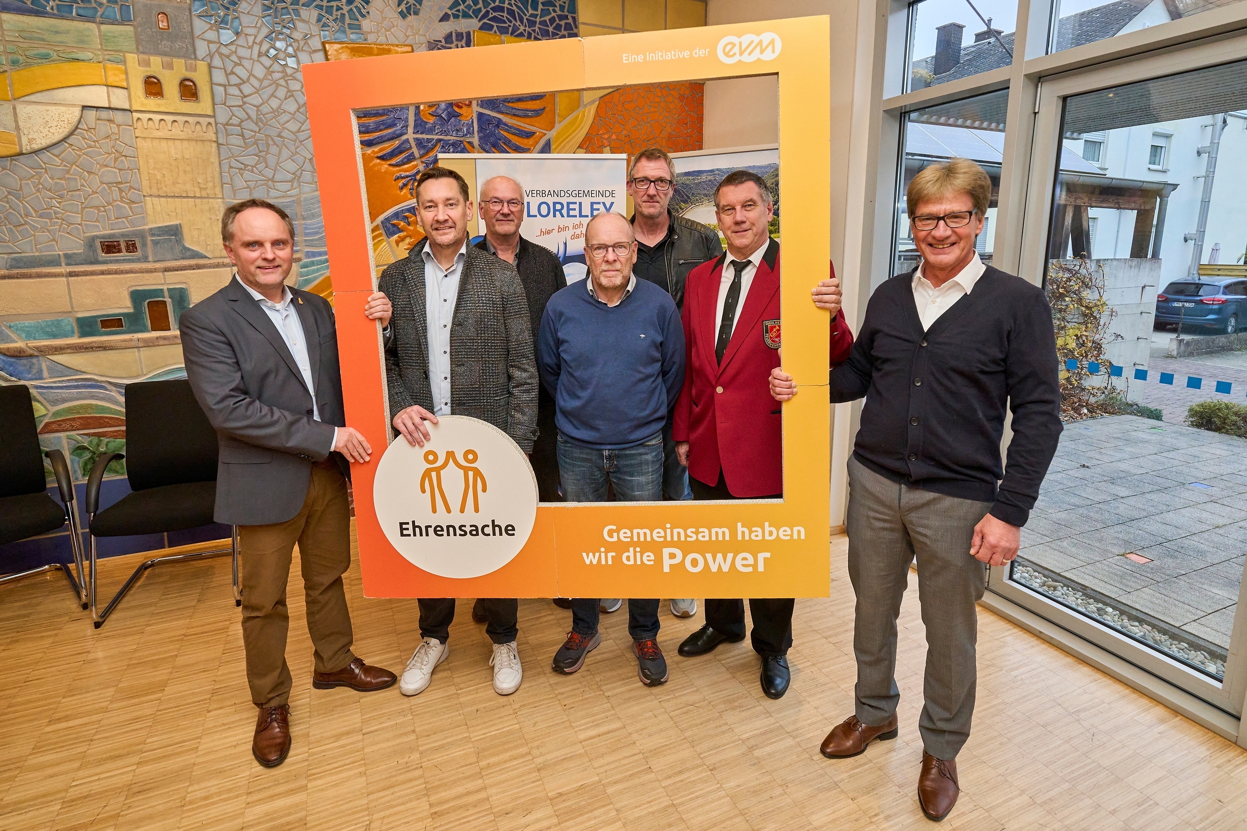 Sieben Männer stehen in einem Raum und halten einen orangenen Rahmen mit der Aufschrift "evm Ehrensache". Sie lächeln freundlich.