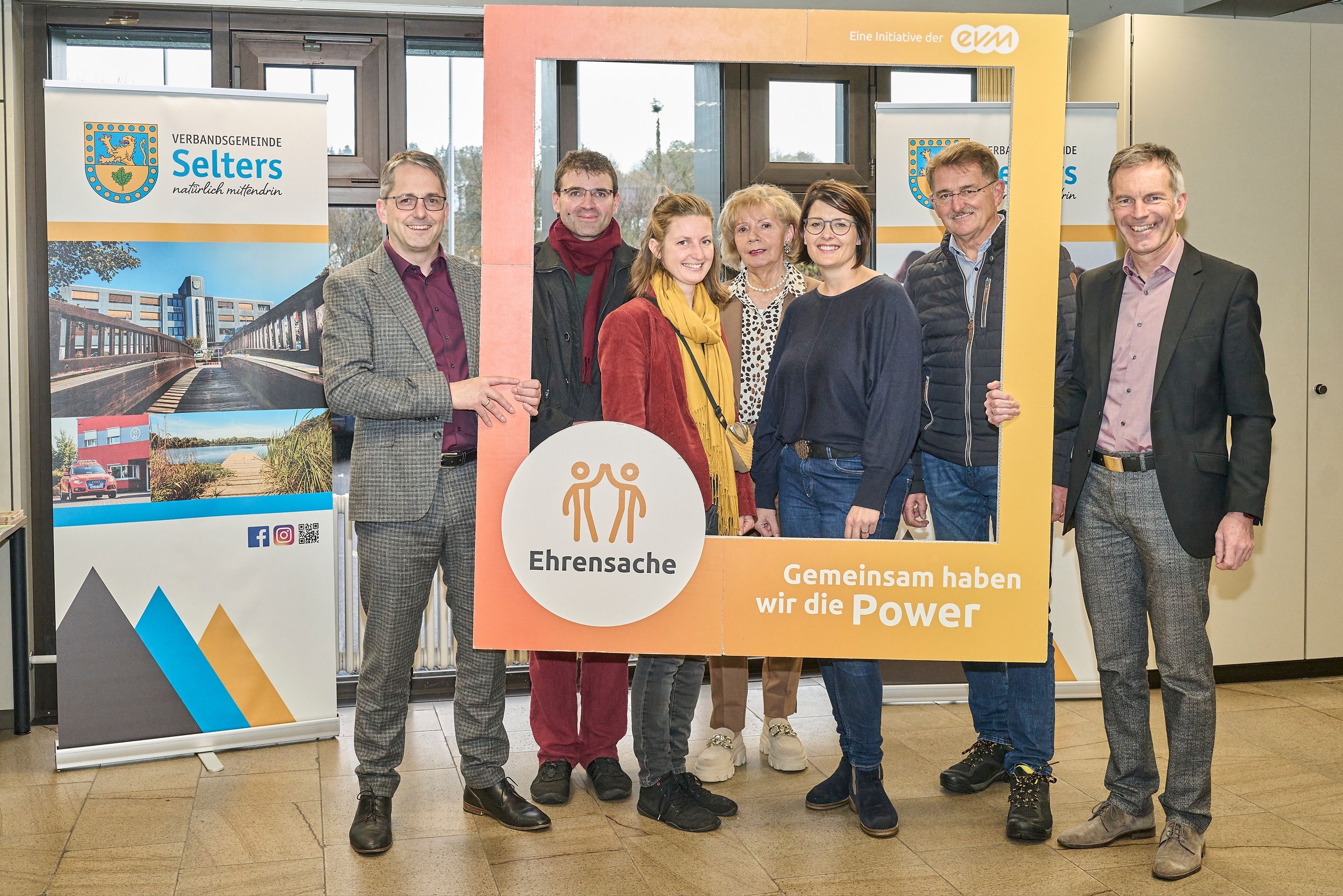 Drei Männer und drei Frauen stehen in einem Raum und halten einen orangenen Rahmen mit der Aufschrift "evm Ehrensache". Sie lächeln freundlich.
