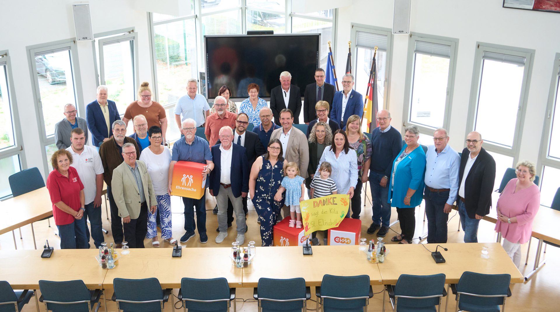 EVM-Ehrensache Weißenturm Eine große Gruppe steht in einem Saal und lächelt freundlich. Eine Person hält einen orangenen Würfel mit Aufschrift "evm Ehrensache".