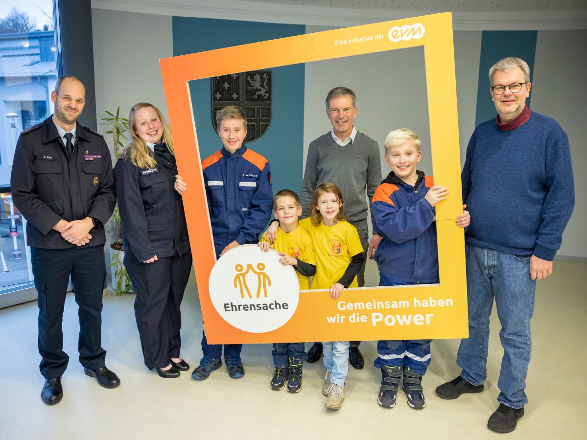 Vier Kinder und vier Erwachsene in Uniformen stehen in einem Raum und halten einen orangenen Rahmen mit der Aufschrift "evm Ehrensache". Sie lächeln freundlich.