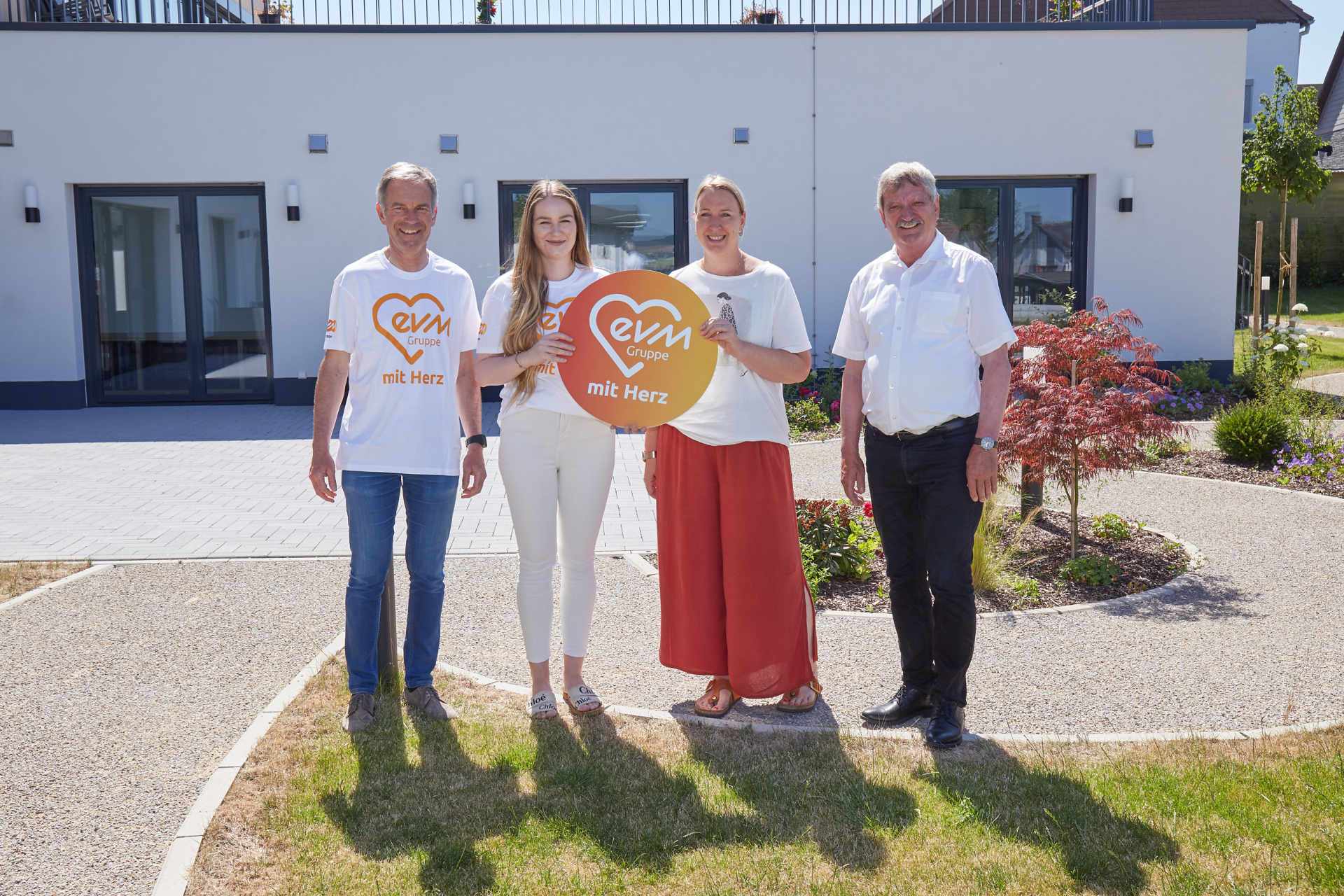 058_012 Zwei Frauen und zwei Männer stehen vor einem Gebäude und lächeln freundlich. Eine der Frauen und einer der Männer tragen ein weißes T-shirt mit orangener Aufschrift "evm mit Herz". Sie halten ein rundes, orangenes evm mit Herz Schild.