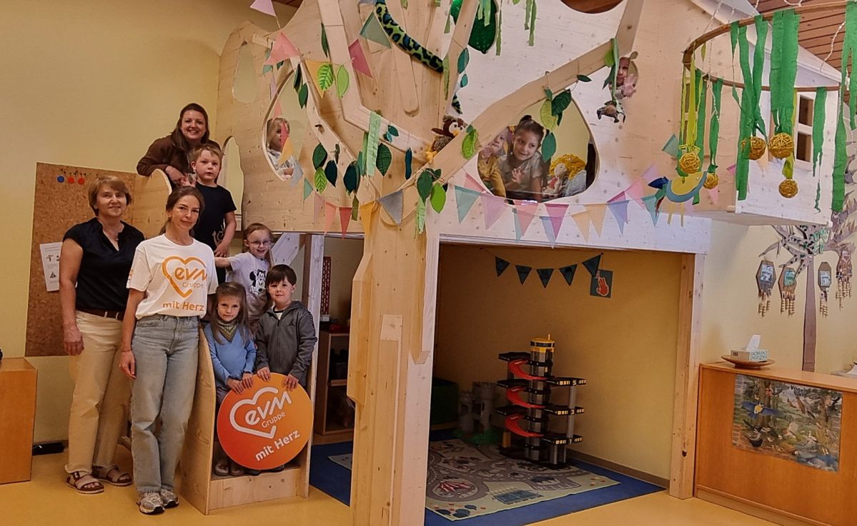 Mehrere Kinder und drei Frauen stehen in einem Kindergarten vor einer Baumhöhle aus hellem Holz. Die Kinder halten ein rundes, orangenes evm mit Herz Schild.