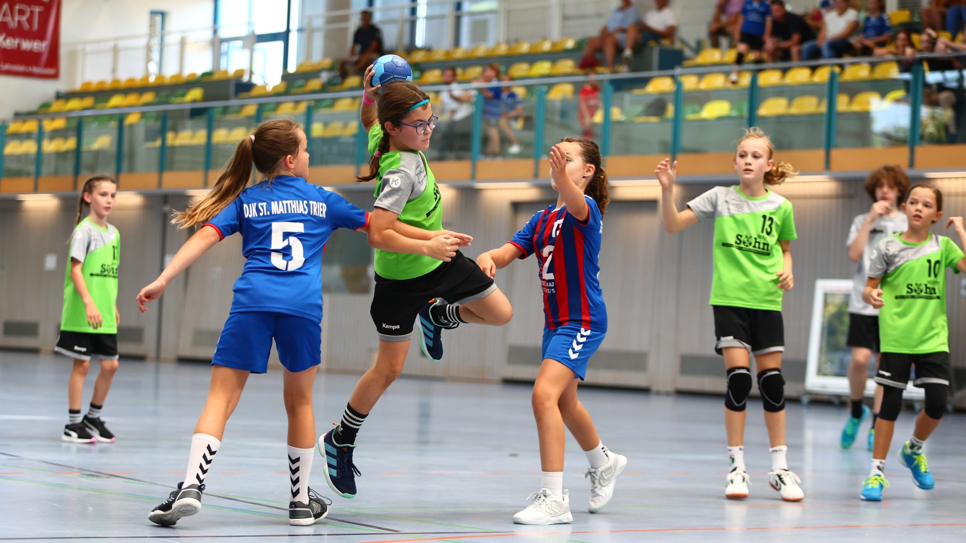 Mehrere Mädchen in Sporttrikots spielen Handball in einer Sporthalle. Im Hintergrund ist Publikum auf Sitzen zu erkennen.