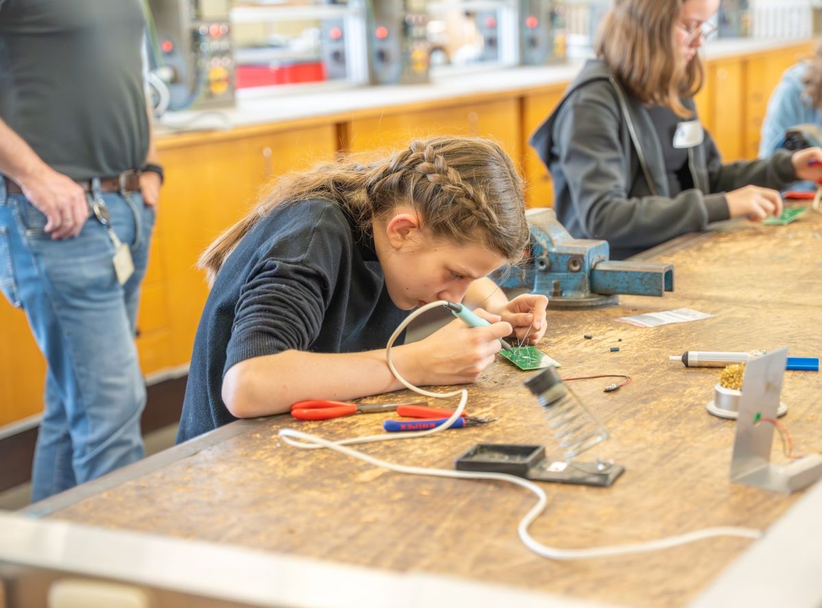 Girls und Boys Day-1 Ein Mädchen mit braunen. geflochtenen Haaren, sitzt in einer Werkstatt und lötet etwas. Um sie herum liegt Werkzeug. Im Hintergrund sind weitere Kinder zu erkennen.