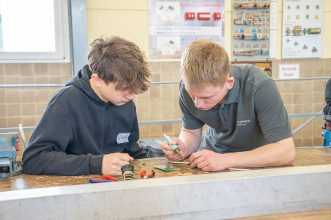 Girls und Boys Day-2 Einem Jungen wird das löten erklärt von einem Azubi. Sie sitzen an einem Tisch in einer Werkstatt. Um sie herum liegt Werkzeug.