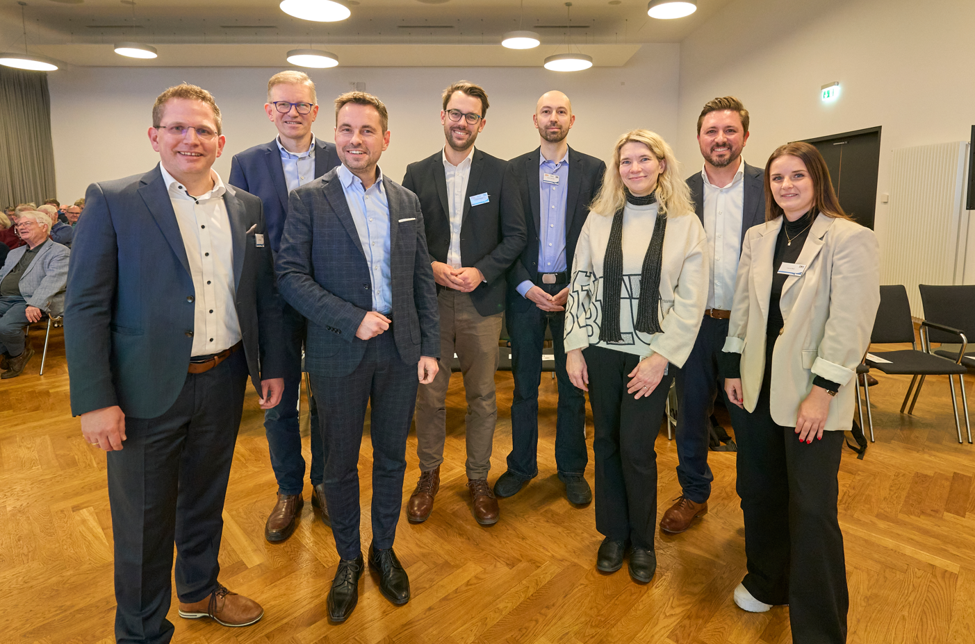 Sechs Männer und zwei Frauen sind professionell gekleidet und lächeln freundlich. Sie stehen in einem Vortragsraum.