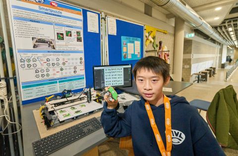 250210SDI1100 Ein Junge steht vor einem Roboter und einem Computer und hält ein Puzzlestück in der Hand.