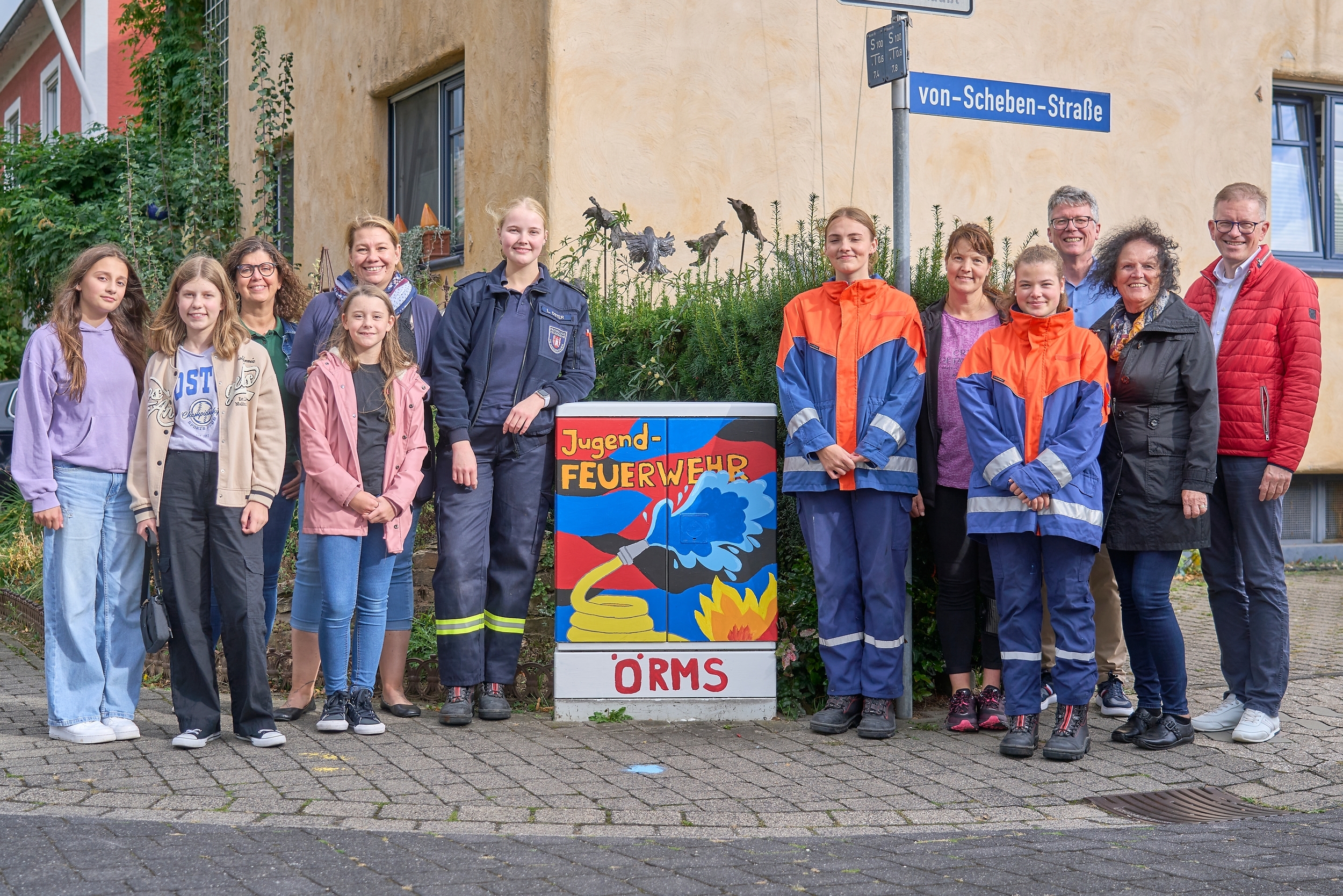 Eine Gruppe von Menschen steht lächelnd vor einem Stromkasten. Zwei tragen Feuerwehruniformen. . Die Umgebung ist eine gepflasterte Straße.
