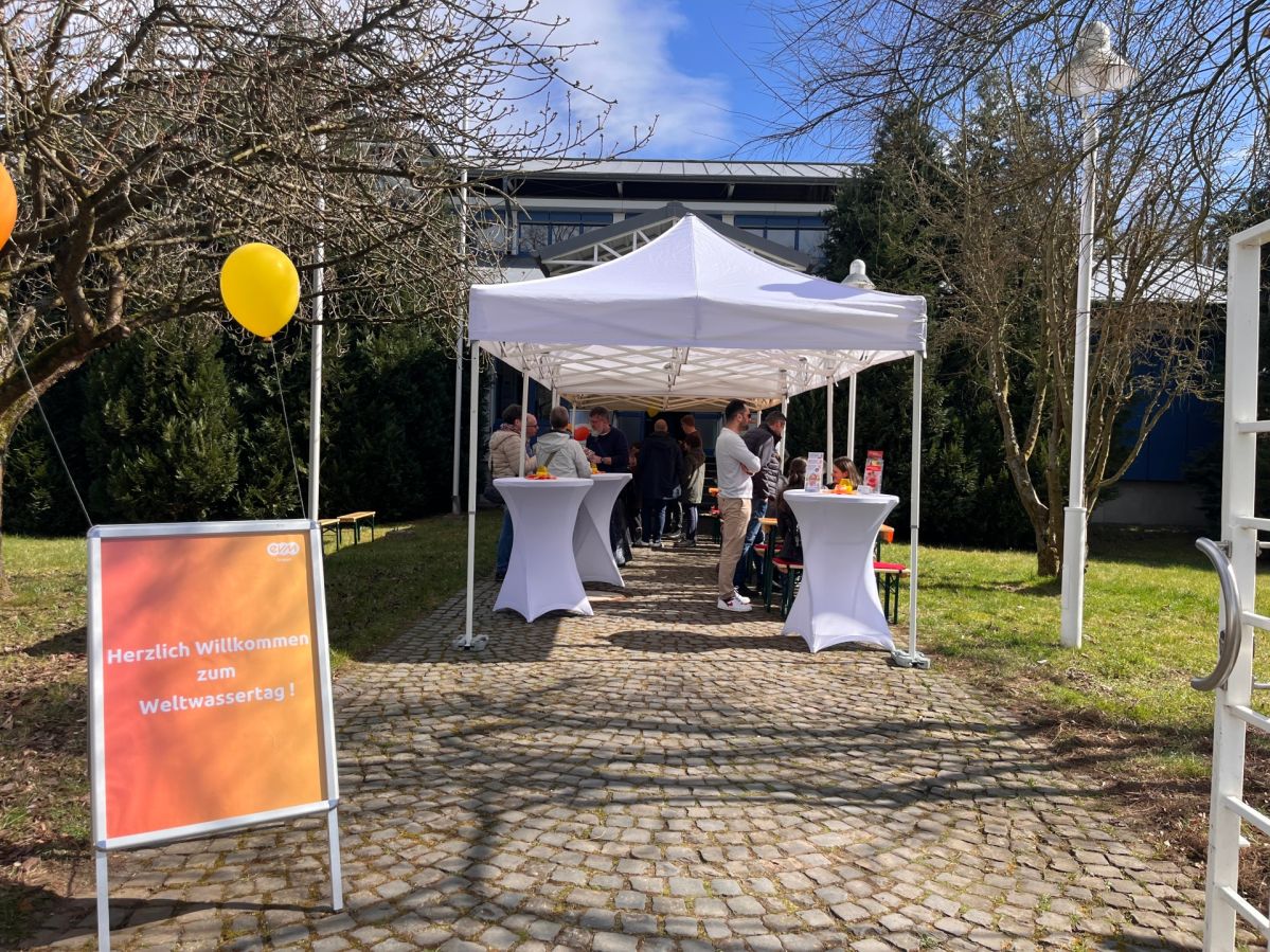Ein weißer Pavillon mit Stehtischen und Ballons steht an einem Gebäudeeingang.  Mehrere Menschen stehen an den Tischen und unterhalten sich. Links steht ein orangener Plakatständer mit Text "Herzlich Willkommen zum Weltwassertag".