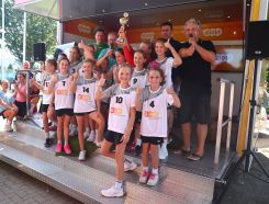 Eine Gruppe von Mädchen in Sportbekleidung posiert mit einem Pokal auf einer Bühne. Im Hintergrund stehen Erwachsene und es gibt ein EVM-Banner. Zuschauer sitzen und stehen am Rand.