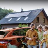 Eine Familie steht neben einem E-Auto dass an einem gelben Ladekabel lädt. Sie stehen vor einem Haus mit einer PV-Anlage.
