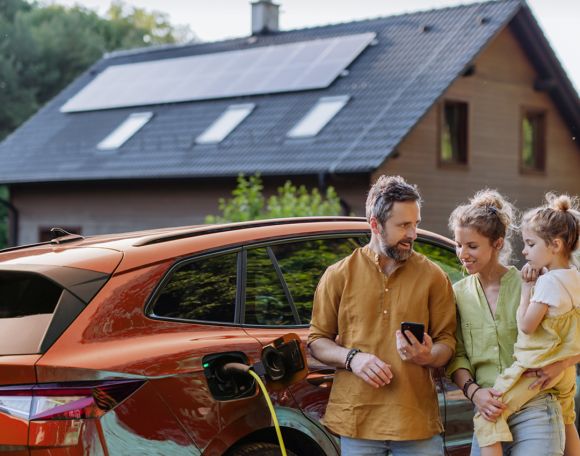 Eine Familie steht neben einem E-Auto dass an einem gelben Ladekabel lädt. Sie stehen vor einem Haus mit einer PV-Anlage.