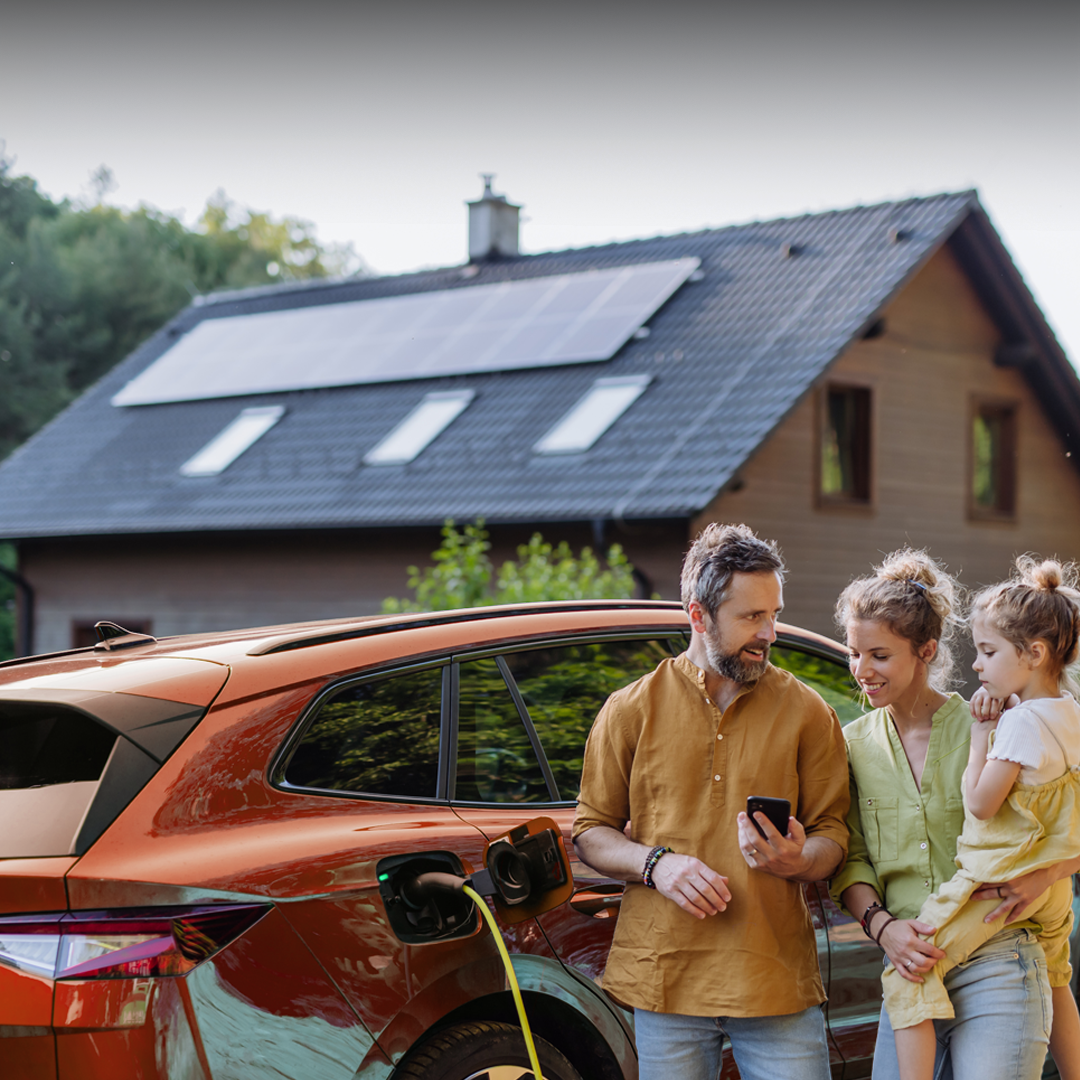 Eine junge Familie steht an ihrem E-Auto. Im Hintergrund ist ein Haus mit Solaranlage auf dem Dach zu sehen