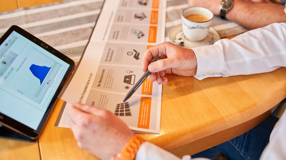 Eine Person sitzt an einem Tisch mit einer anderen Person. Vor ihnen steht ein Tablet und sie zeigen auf einen Fyler.