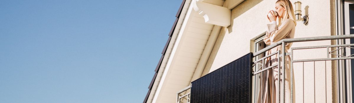 Ein Solarpanel ist an einem Balkongelände montiert. Eine Frau steht auf dem Balkon und trinkt aus einem Becher.